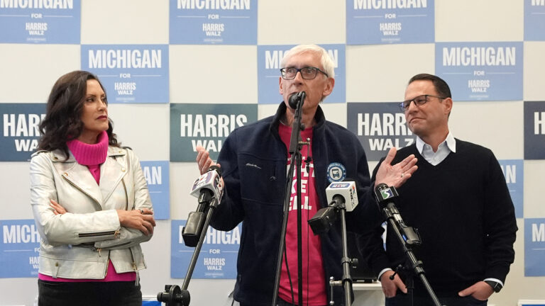 Tony Evers gestures with both hands while standing and speaking into multiple microphones with the flags of different media organizations mounted on stands, with Gretchen Whitmer and Josh Shapiro standing on either side of him, in a room with political posters reading Michigan for Harris Walz displayed in a checkerboard pattern on the wall behind them.