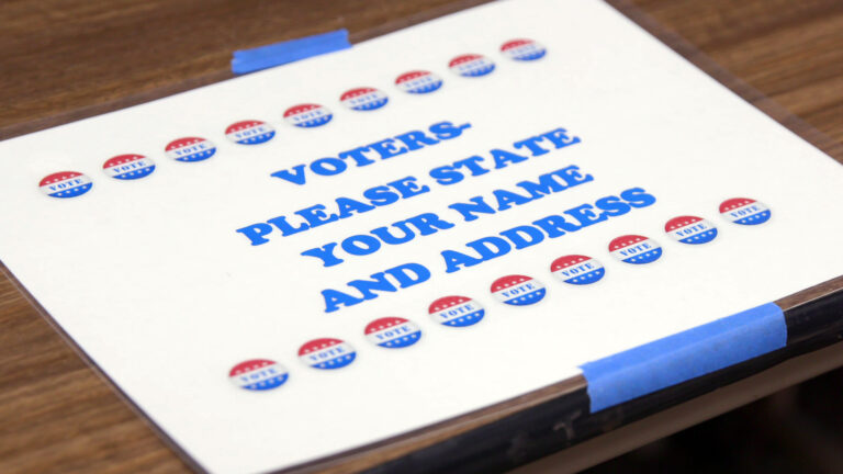 A printed sheet of paper with two rows of graphics in the shape of buttons reading Vote with the text Voters - Please State Your Name and Address is encased in a plastic sheet protector and attached with painters tape to the wood laminate surface of a table.