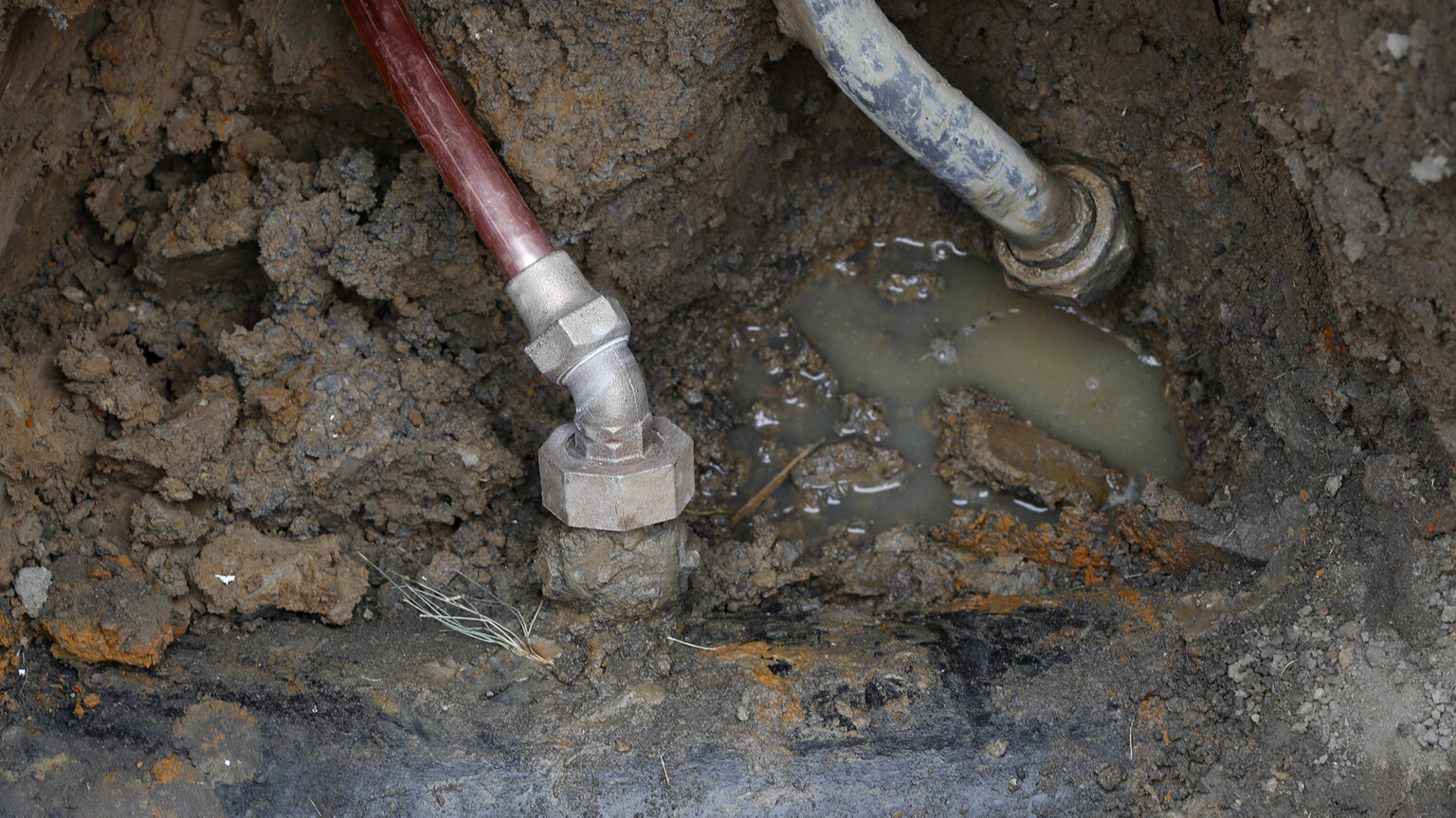 Two narrow gauge metal pipes and one wider metal pipe are half buried in dirt, with small puddles of water in  several indentations in the soil.