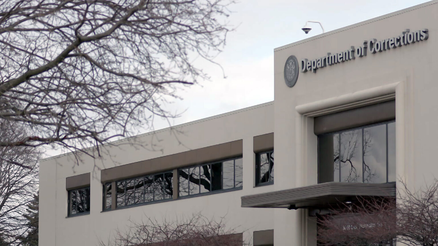 Leafless trees stand in front of and to the side of a two-story concrete-masonry building with a central entrance topped by signage with the seal of the state of Wisconsin and the words Department of Corrections.