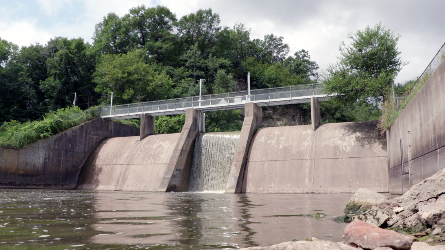 Water tumbles over the edge of a channel of a concrete dam on a river, with a fenced walkway spanning its top and concrete retaining walls on both sides, with foamy water and shore rocks in the foreground and trees in the background.