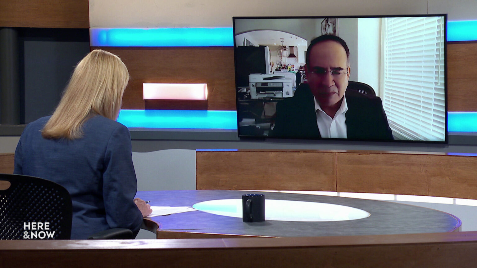 Frederica Freyberg sits at a desk on the Here & Now set and faces a video monitor showing an image of Jorge Franco.