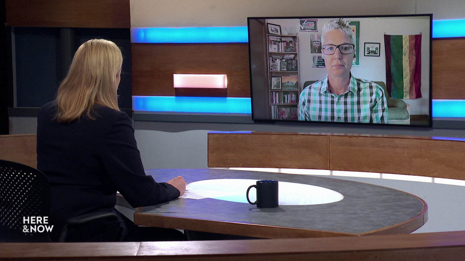 Frederica Freyberg sits at a desk on the Here & Now set and faces a video monitor showing an image of Abigail Swetz.