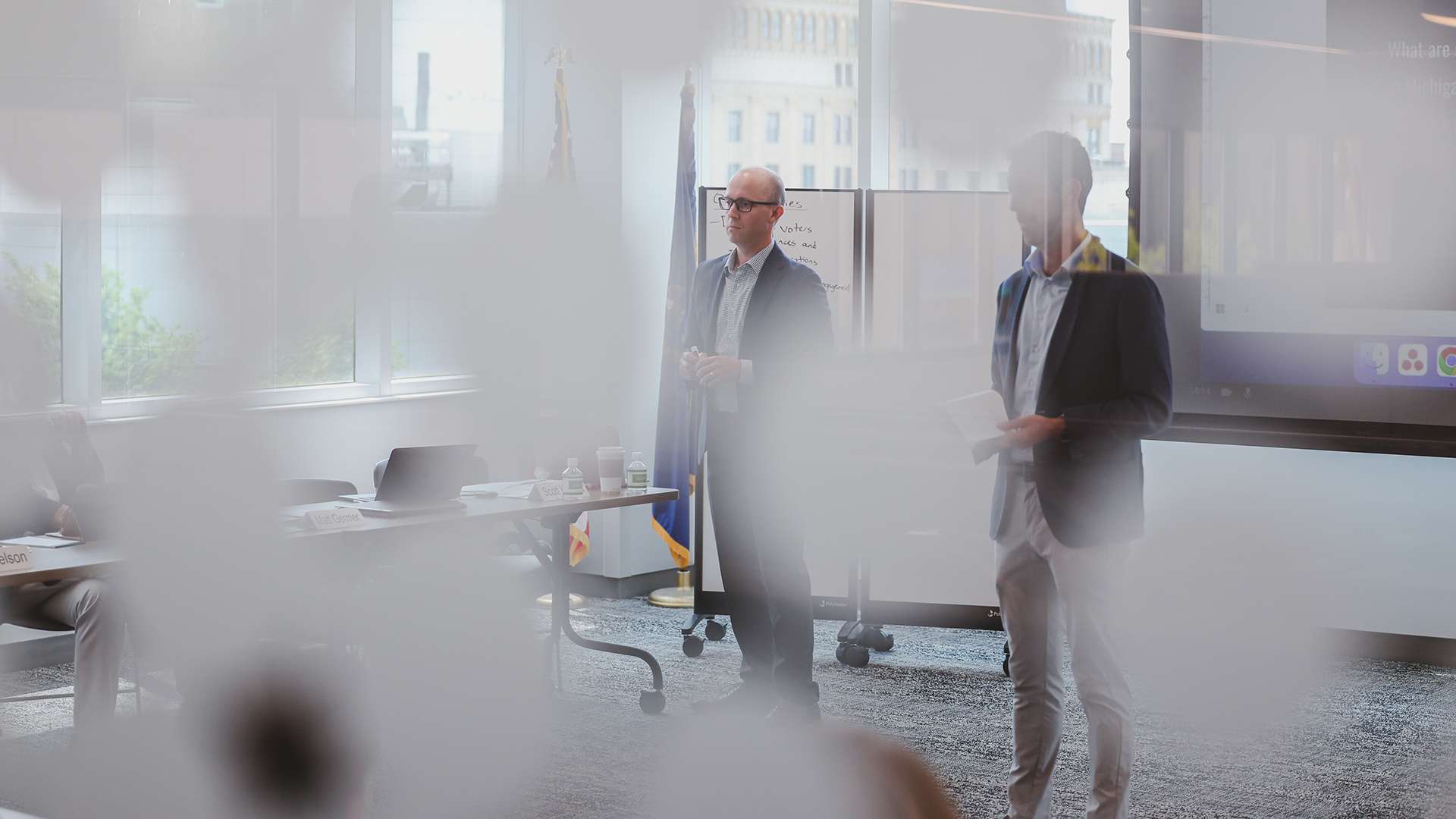 Matt Germer and Scott Warren are seen through out-of-focus circular window decals while standing inside a room with a large wall-mounted monitor, a rolling table, a rolling whiteboard and large plate-glass windows with neighboring buildings visible outside.