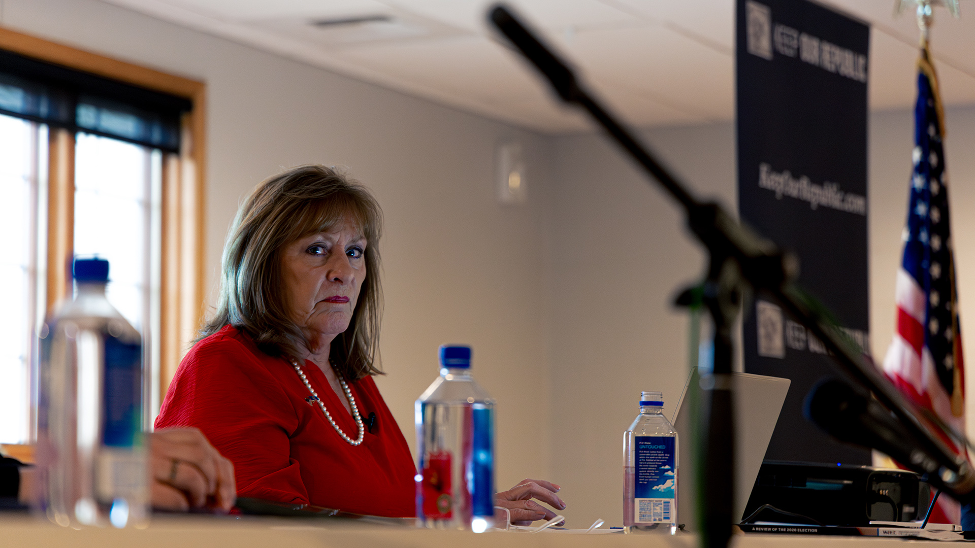 Kathy Bernier sits at a table with multiple plastic bottles of water and an open laptop computer on its surface, with an out-of-focus window and U.S. flag in the background, and an out-of-focus microphone stand in the foreground.