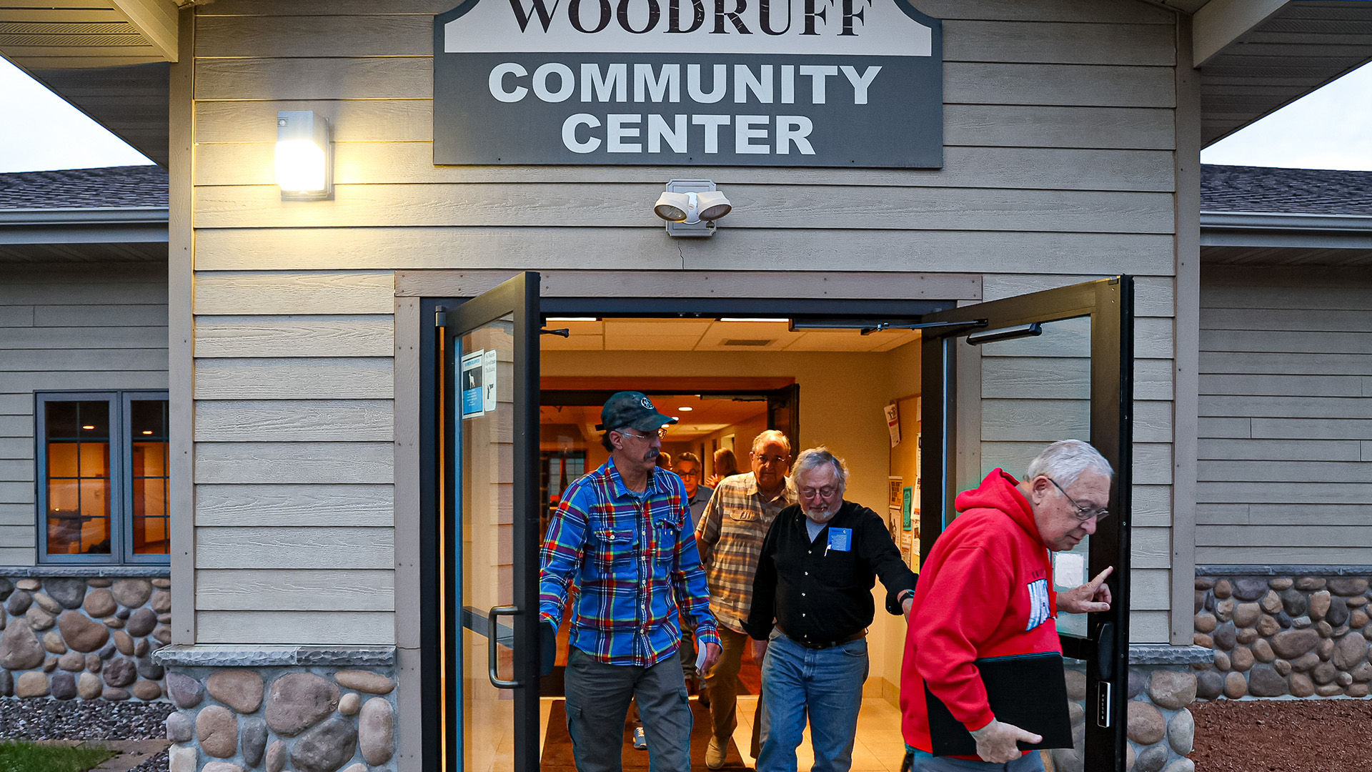 Multiple people walk between a pair of open metal and glass double doors, exiting a building with stone and wood siding, an illuminated exterior light and a sign reading "Woodruff Community Center."