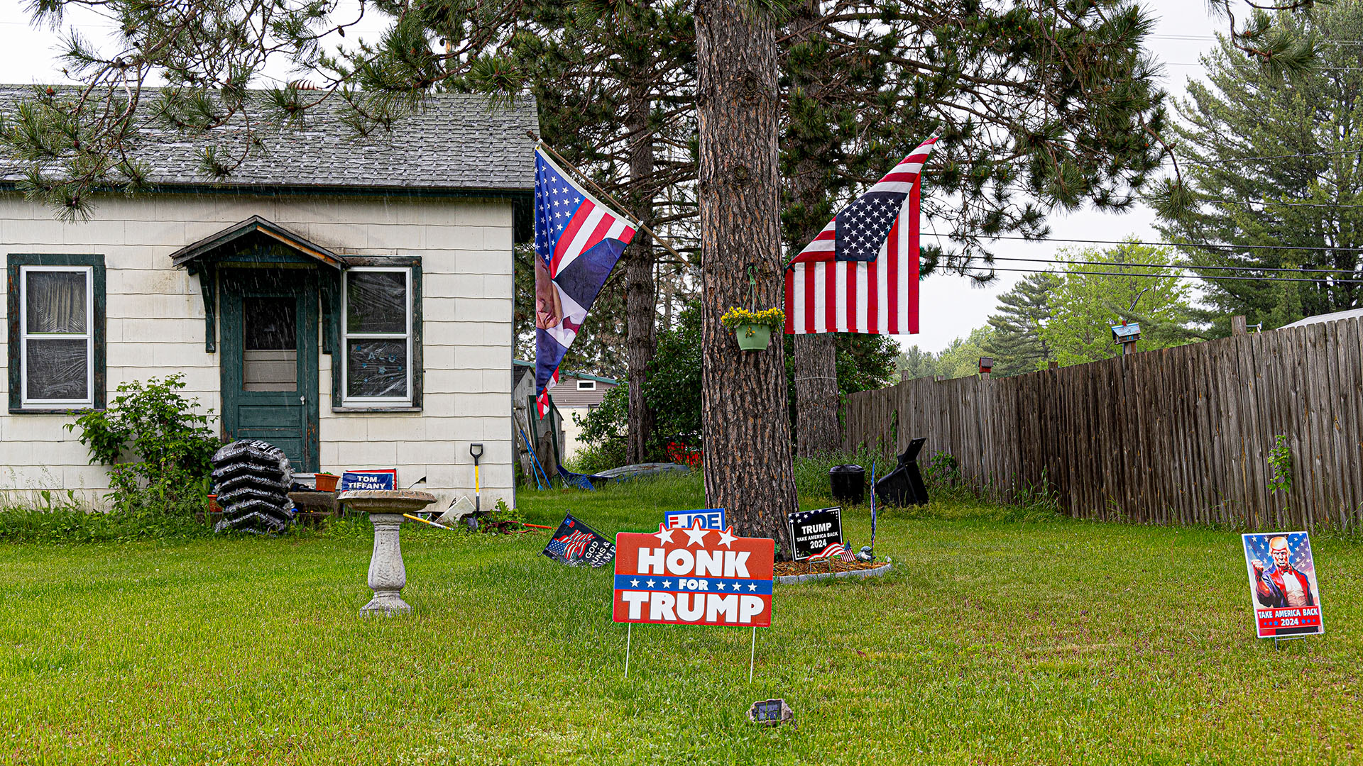 Multiple political signs, including one reading "Honk for Trump," are placed in a lawn in front of a tree with an upside-down U.S. flag and another flag showing Donald Trump superimposed over stars and stripes mounted to its trunk, with a house, wood fence and more trees in the background.