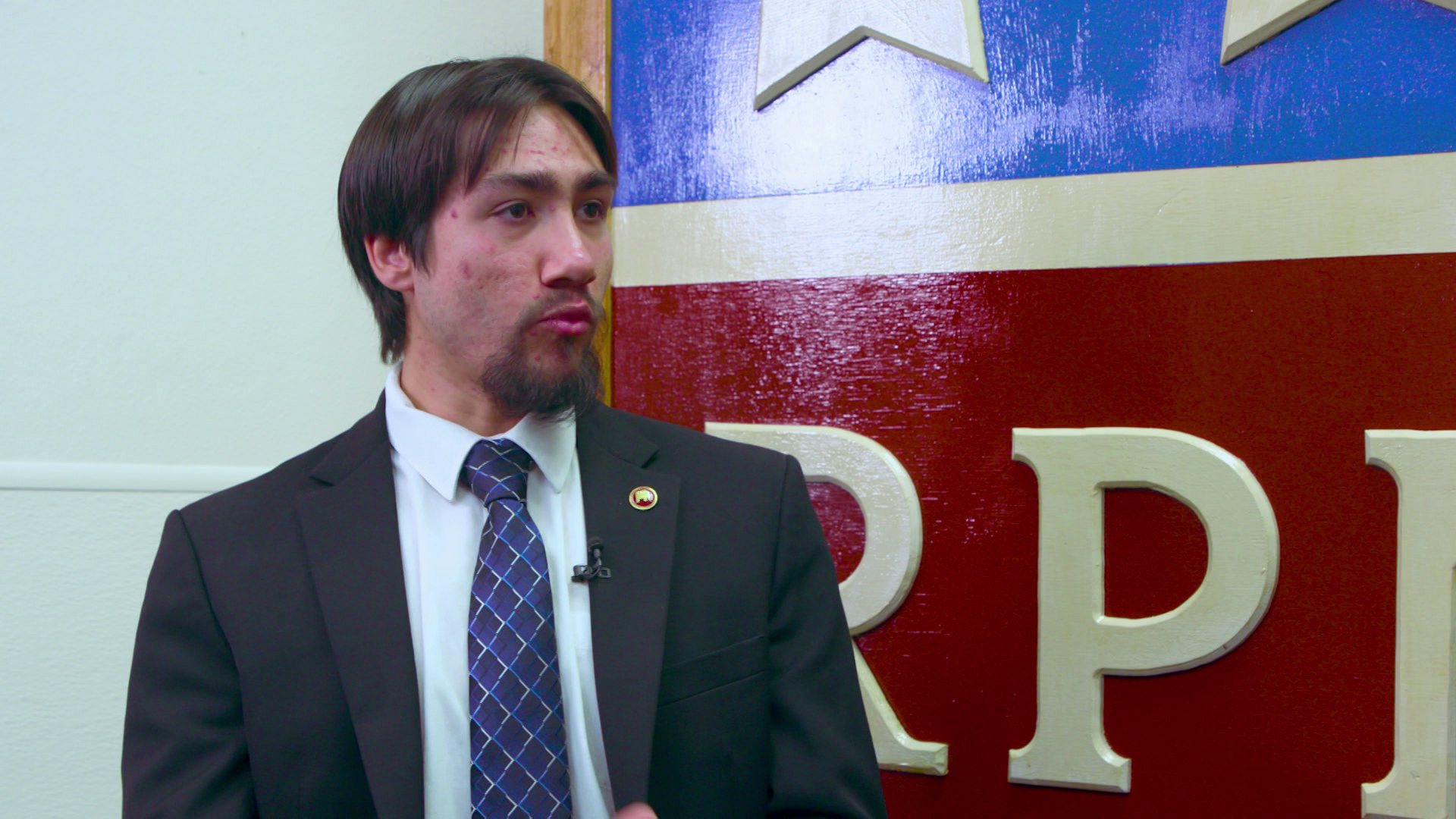 Hilario Deleon speaks while sitting indoors in front of a large, painted wooden sign with letters and stars behind him.