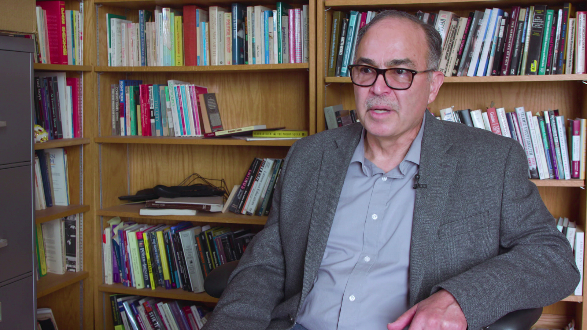 Benjamin Marquez speaks while sitting indoors with wood bookshelves lined with books and other items in the background.