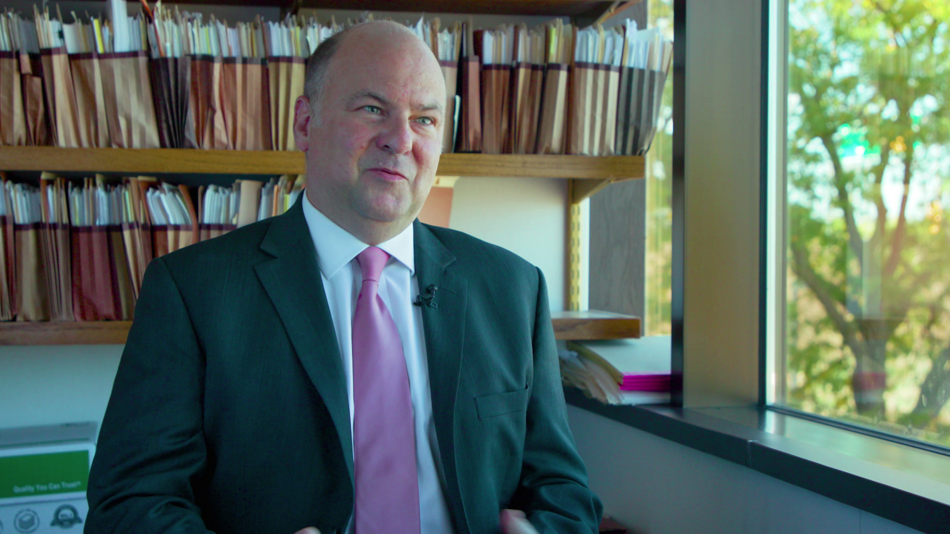 Davorin Odrcic speaks while sitting indoors with expandable file folders containing papers lining shelves mounted on the wall and a window with trees outside visible in the background.