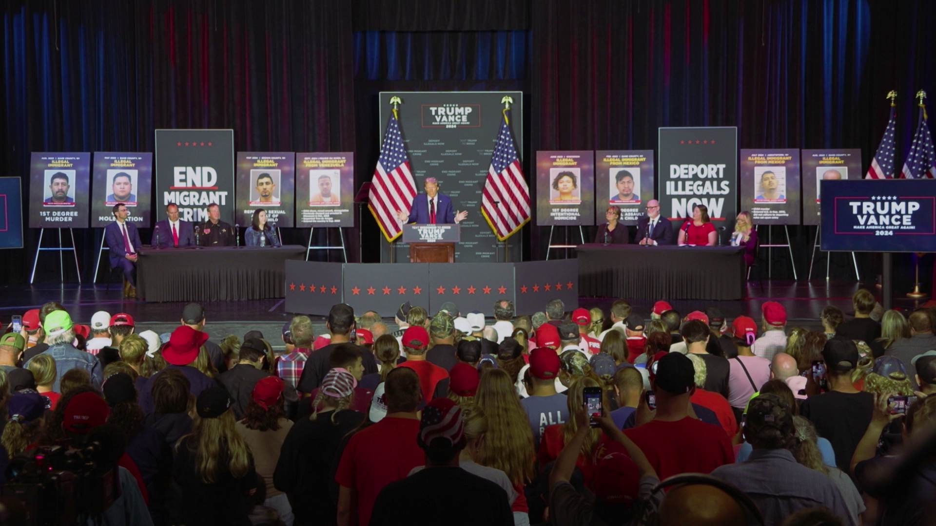 Donald Trump speaks while standing behind a podium on a temporary stage with people seated at tables on either side of him, and with U.S. flags and printed signs on tripods showing photos of immigrants convicted of crimes and political slogans, with rows of seated people facing him in the foreground.