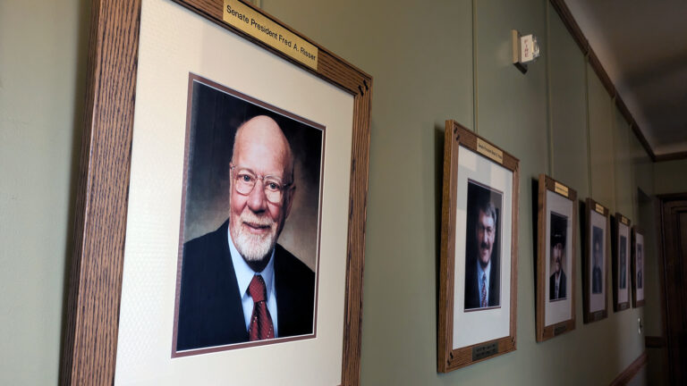 A portrait of Fred Risser is displayed in a matted frame with a brass label at its top reading Senate President Fred A. Risser, with five other similarly framed and labeled portraits displayed in a row along a wall to its side.