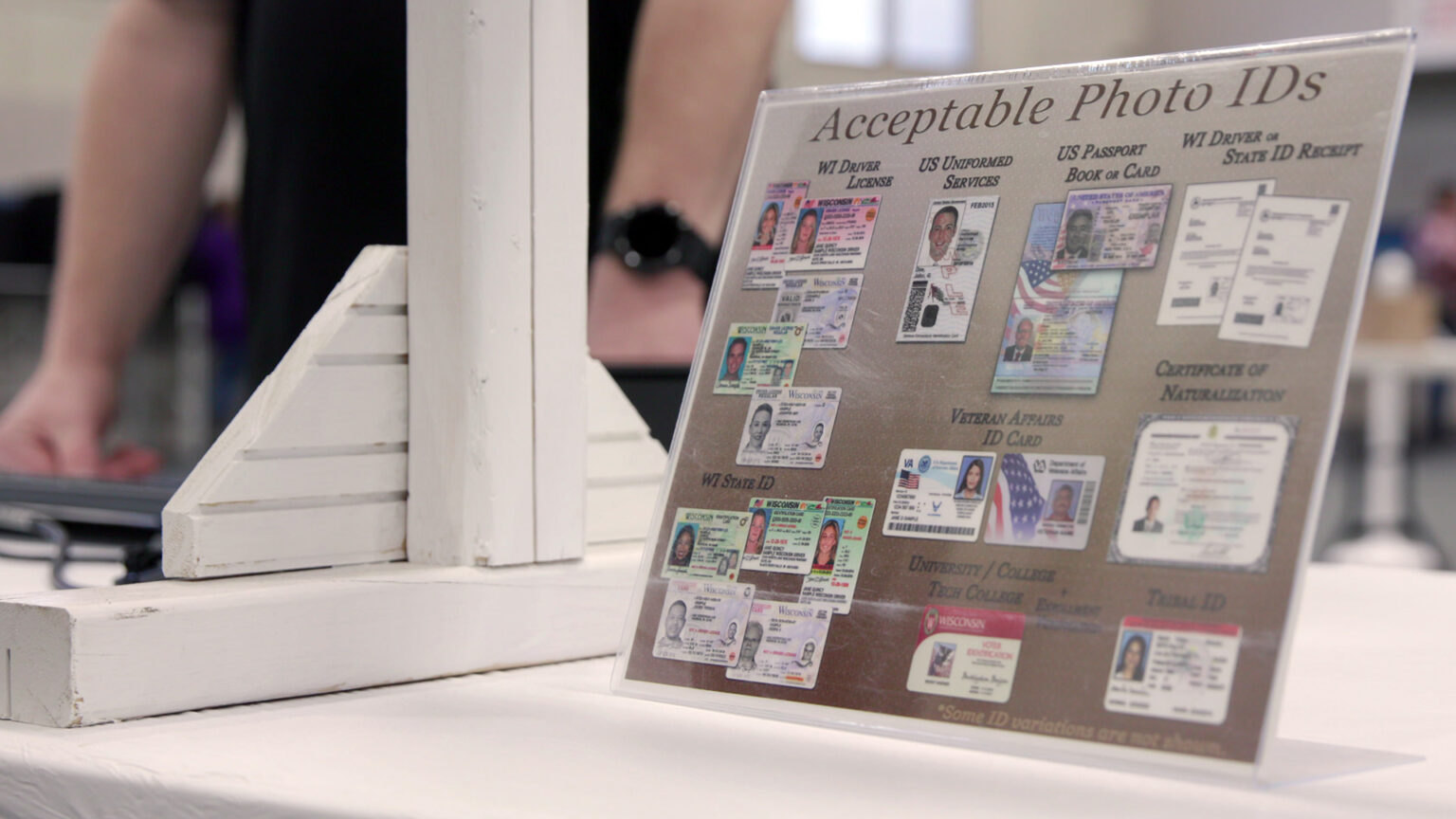 A printed paper sign with the title Acceptable Photo IDs and examples of different forms of photo identification is encased in a plastic display stand atop a table next to painted wood stand, with an out-of-focus person standing in the background.