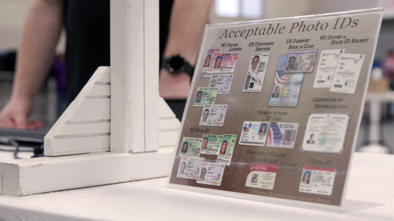 A printed paper sign with the title Acceptable Photo IDs and examples of different forms of photo identification is encased in a plastic display stand atop a table next to painted wood stand, with an out-of-focus person standing in the background.