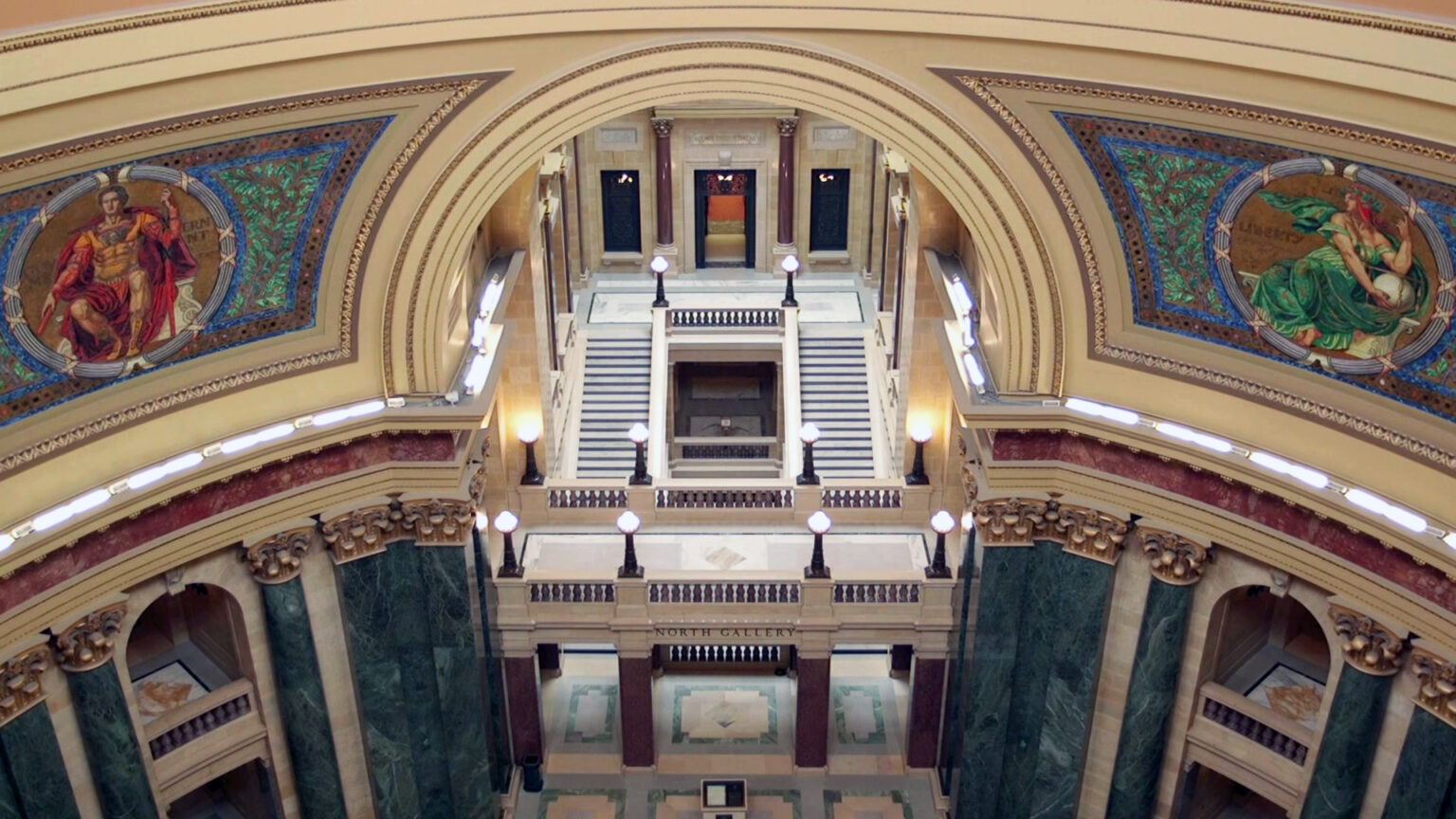 Two mosaics depicting Classical figures — one holding a rod and labeled Government and the other holding a globe and labeled Liberty — are seen from above in two quadrants of the interior walls of a rotunda inside a building with marble pillars and other masonry in a Neoclassical design, with stairs and walkways connecting levels and rooms in one wing.