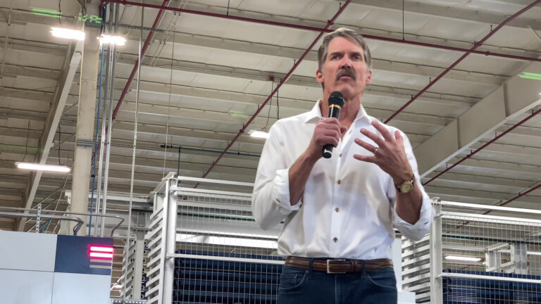 Eric Hovde speaks and holds a wireless microphone in his right hand and gestures with his left hand while standing in a warehouse with metal racks in the background and a high ceiling with fluorescent lighting and a sprinkler system.