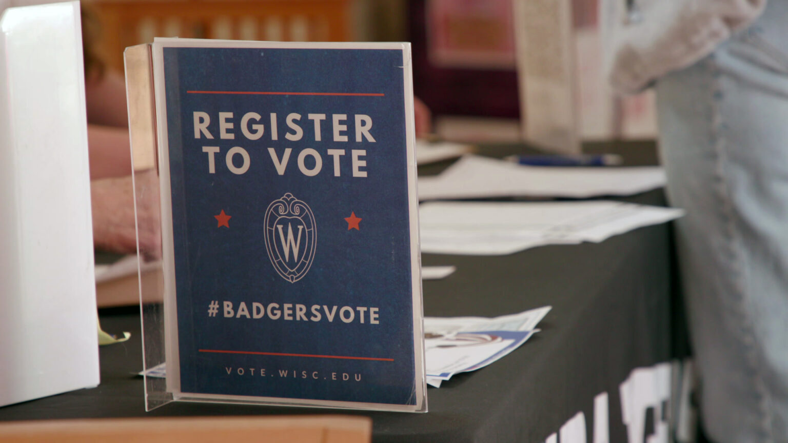 A printed paper sign showing the UW-Madison W Crest and the words Register to Vote is encased in a transparent plastic stand set on a surface of a table with papers on its surface and a standing person on one side facing a seated person on another side in the background.