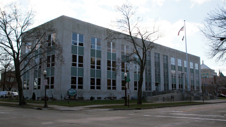 Leafless trees, a flagpole with the U.S. and another flag, and street light poles stand in front of a multi-story masonry building with large glass windows, with other buildings in the background and an intersection of two streets in the foreground.