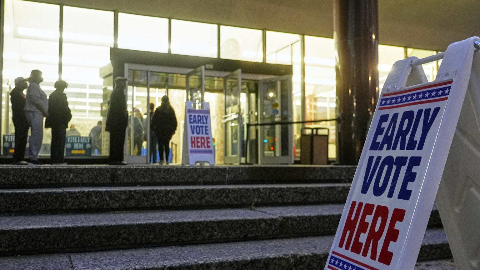 A plastic sandwich board sign with the words Early Vote Here stands at the bottom of a short set of marble steps , with an identical sign standing in front of open glass double doors at the entrance of a building with a glass wall and an exterior pillar as people stand in a line outside and going inside.