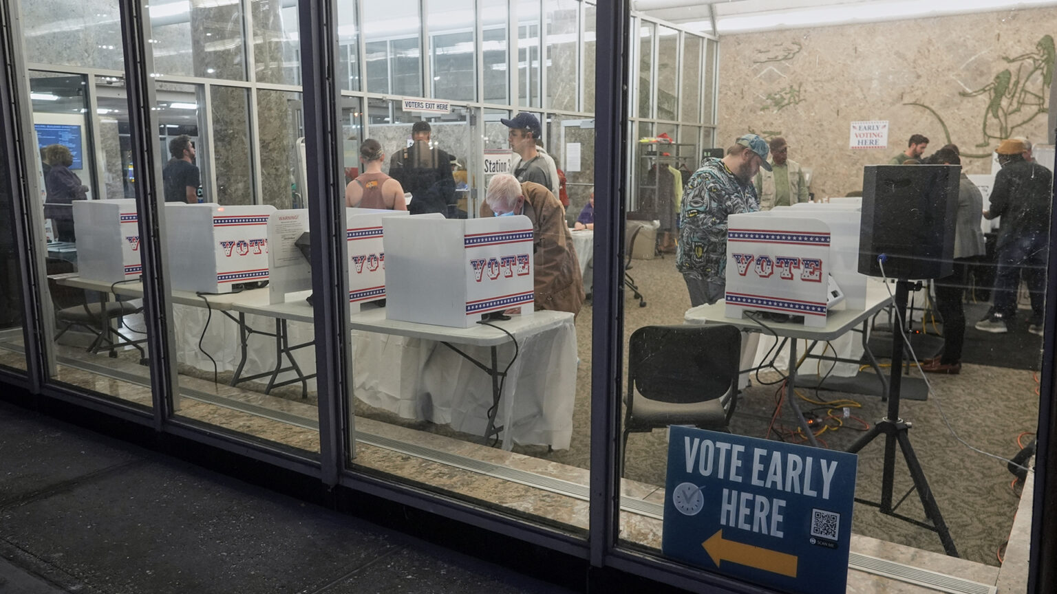 Multiple people stand in front of collapsible three-sided privacy screens with Vote printed on the side set on top of folding tables while other people stand and sit in other locations in a room with a speaker mounted on a tripod, low pile carpet, marble walls and fluorescent lights, as seen from outside through large plate-glass windows with a printed political sign showing an arrow, a QR code and the words Vote Early Here standing at the base of one window.