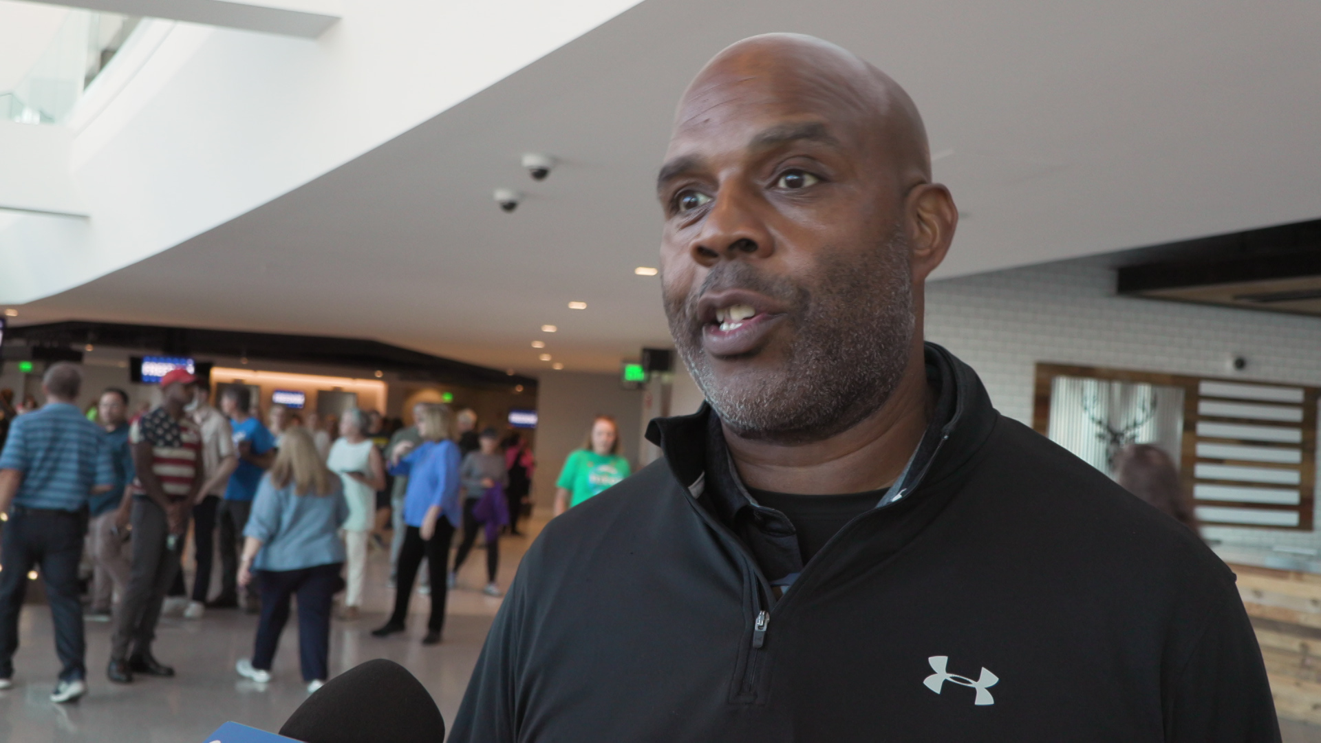 Terrence Burkett speaks into a microphone while standing indoors with people walking in the background.