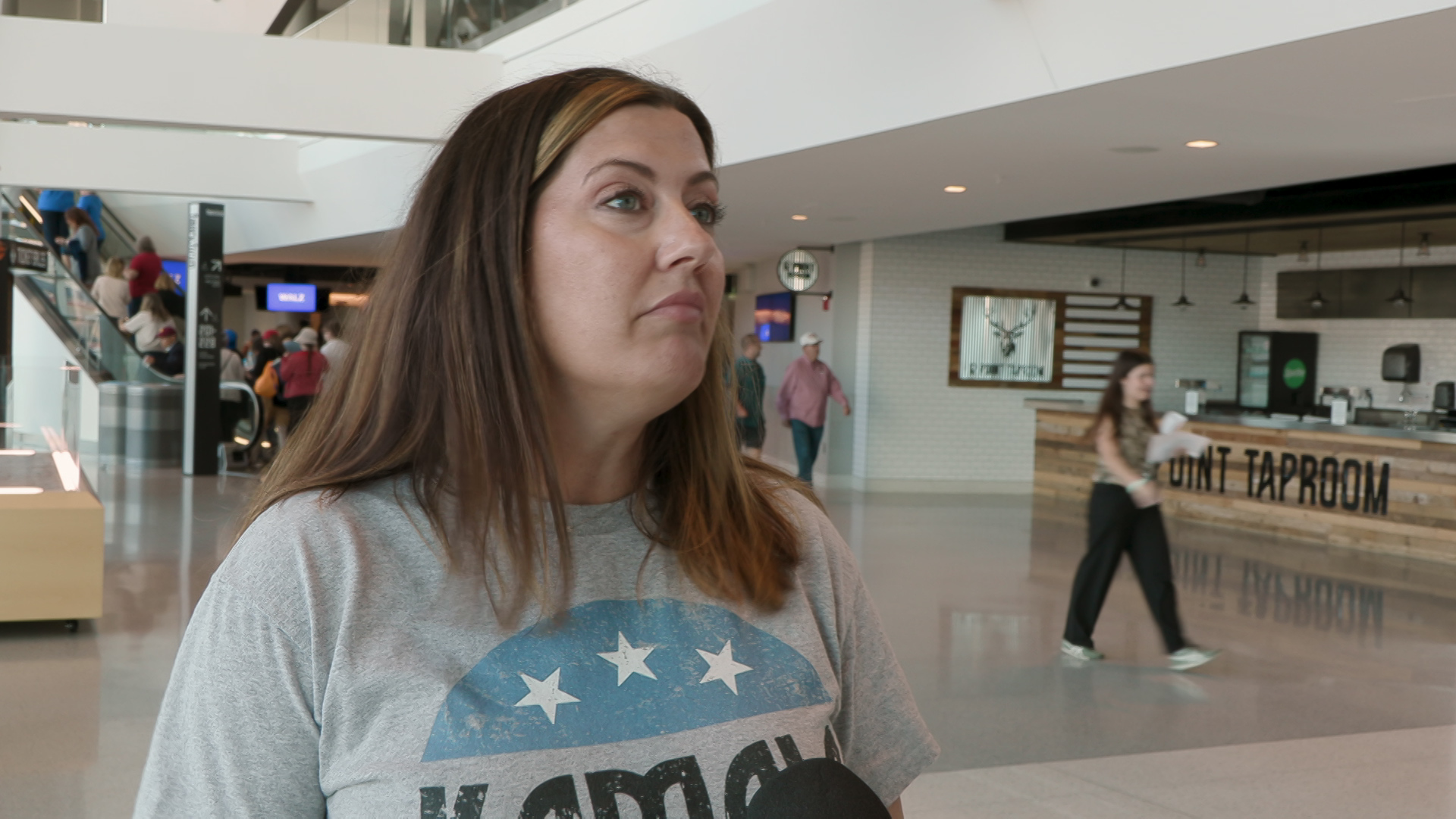 Jessica Cabrera speaks into a microphone while standing indoors with people walking in the background.