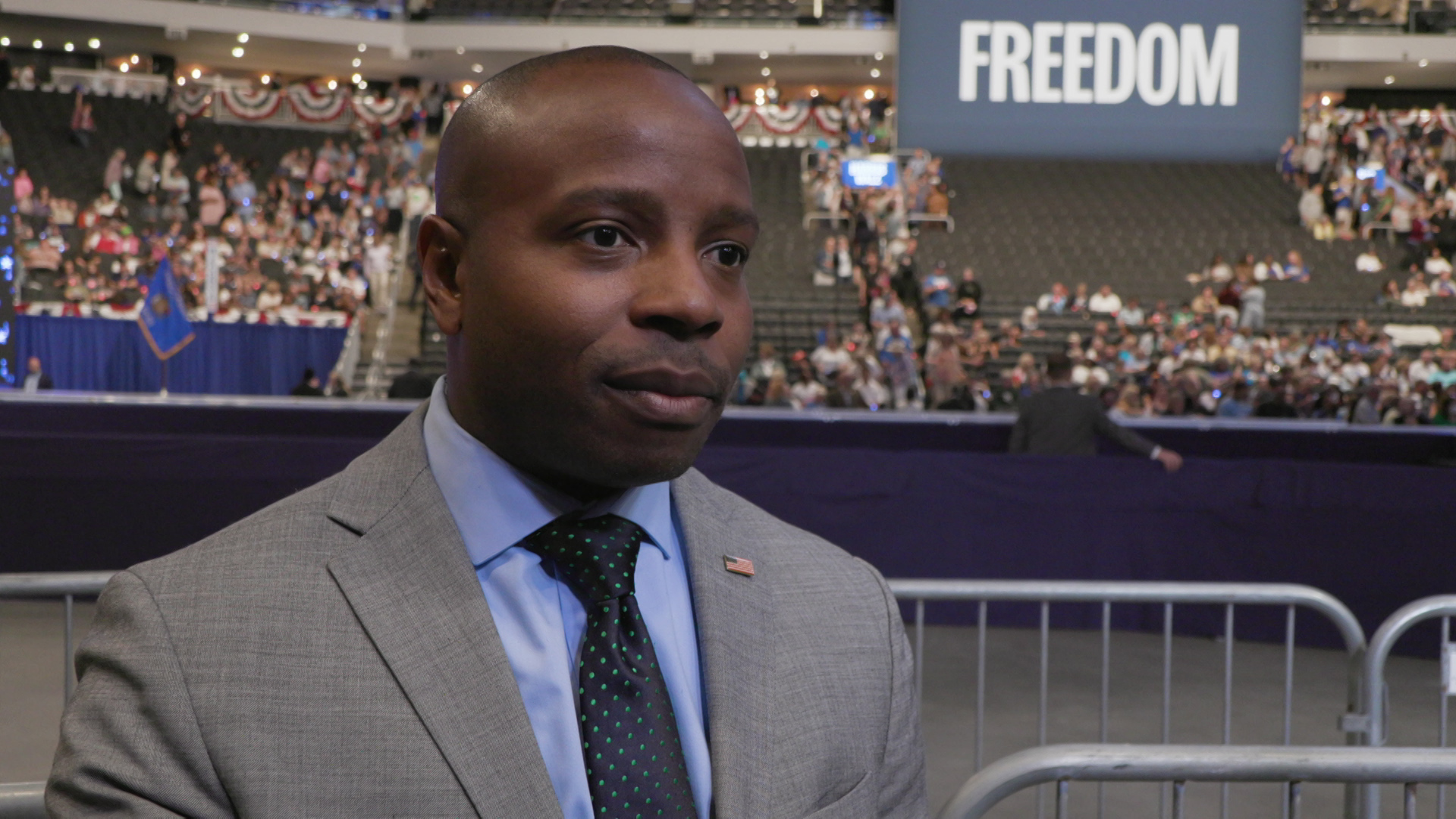 Cavalier Johnson speaks while standing on the floor of an arena with out of focus people sitting in ascending rows of seats, with red, white and blue bunting displayed on railings next to an electronic sign reading "Freedom."