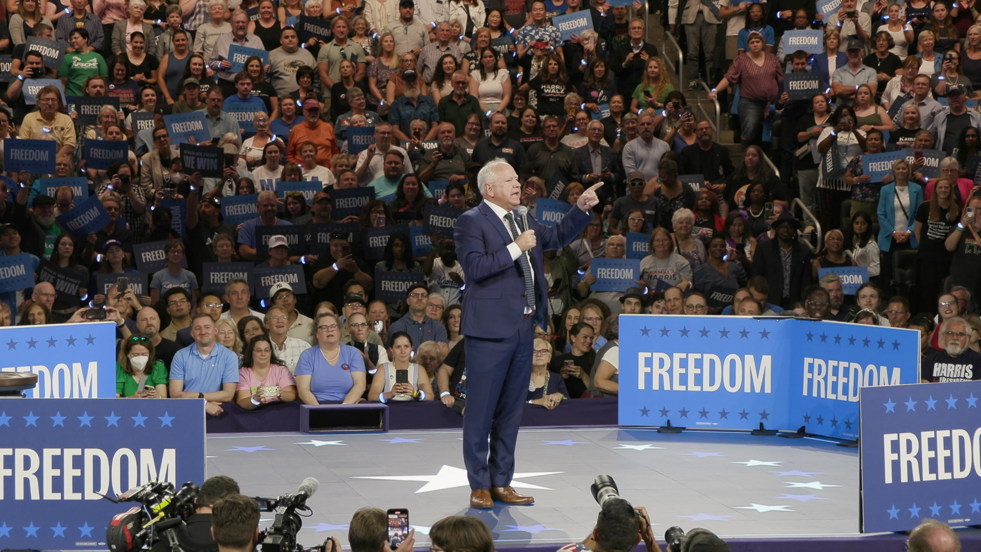 Tim Walz holds a microphone and speaks while standing on a stage with an audience of people in the background holding signs reading "Freedom" and a group of photographers in the foreground.