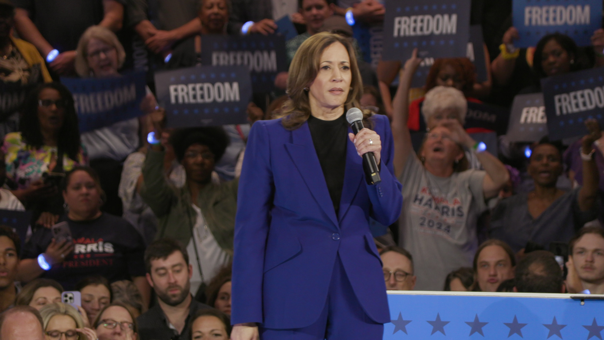 Kamala Harris holds a microphone and speaks while standing in front of an audience of people holding signs reading "Freedom."