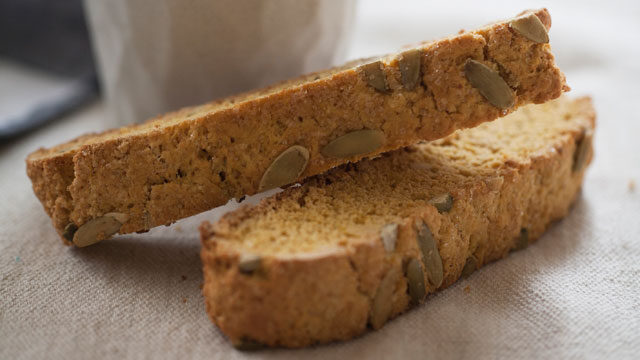 A glamor shot of one biscotti with pumpkin seeds angled on top of another, a mug blurred in the background.