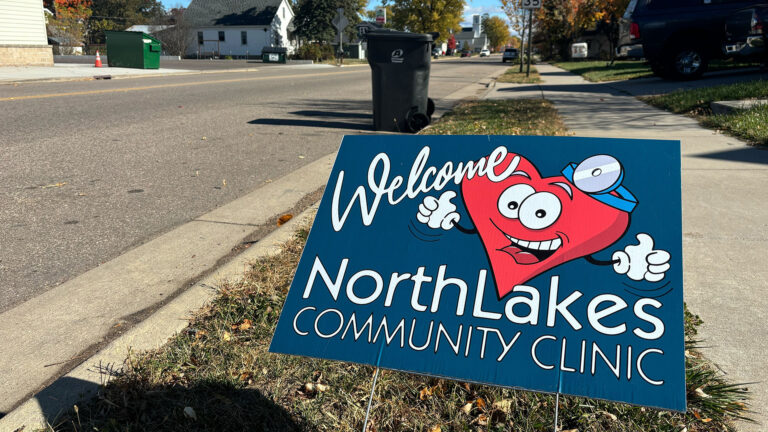 A cardboard yard sign with an illustration of an anthropomorphic heart wearing a head mirror and the words Welcome NorthLakes Community Clinic stands in a grass median between a street and sidewalk, with a lidded plastic trash can, parked vehicles, buildings and trees in the background.