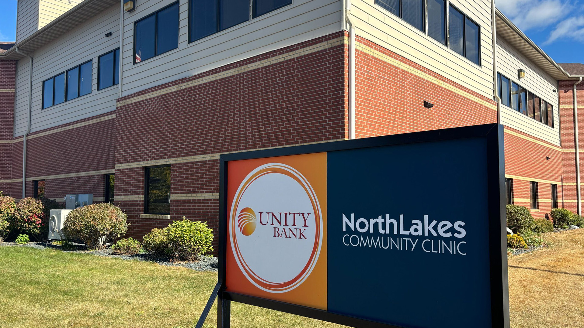 A sign showing the wordmark for Unity Bank and the words "NorthLakes Community Clinic" stands in front of a multi-story brick building with rows of windows on an upper floor and with landscaped bushes and plants at its base.