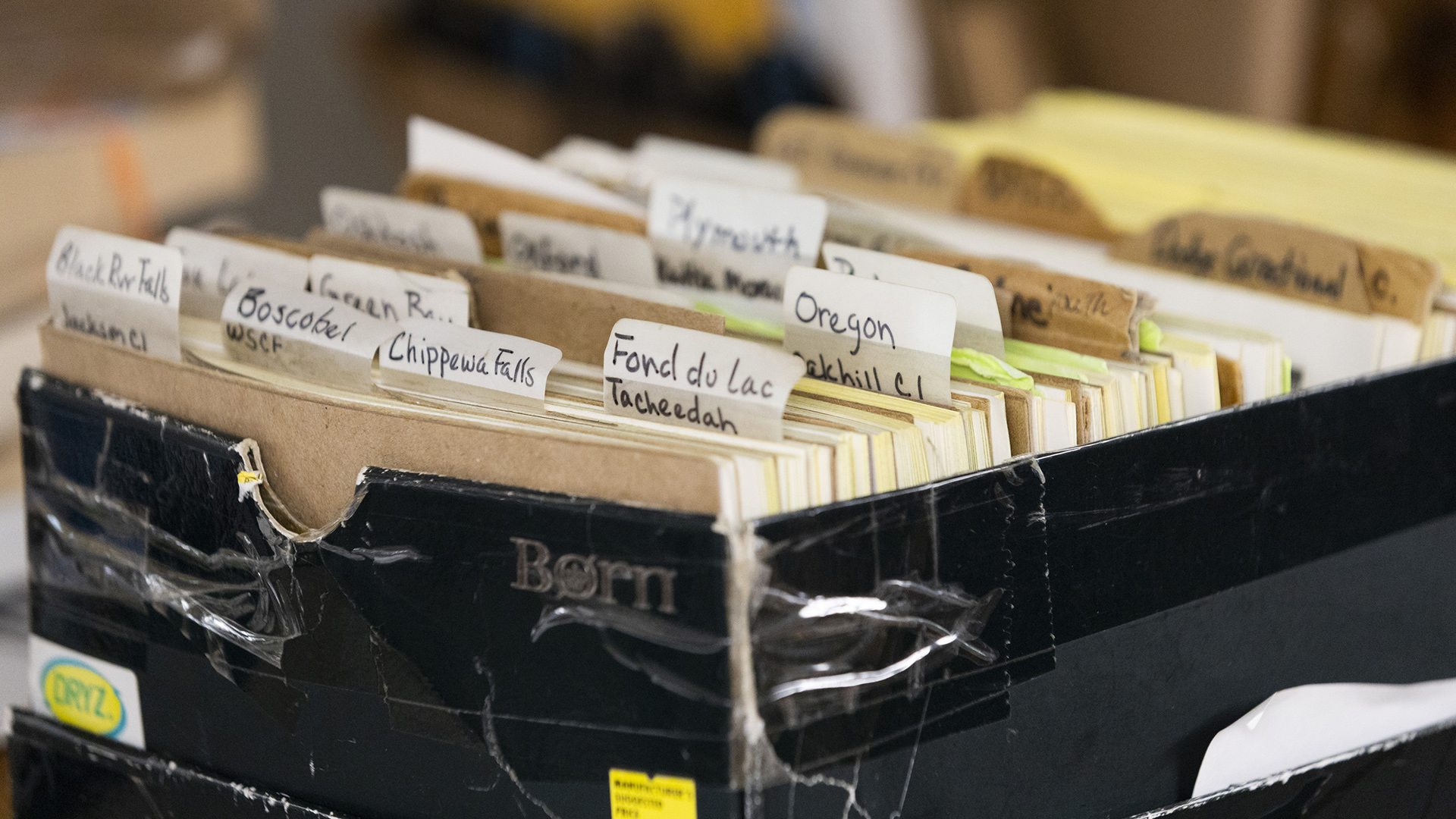 A cardboard box with multiple tape repairs is filled with files with labels that read" "Black River Falls," "Boscobel," "Chippewa Falls," "Fond du Lac Tacheedah," and "Oregon Oakhill CI," among others.