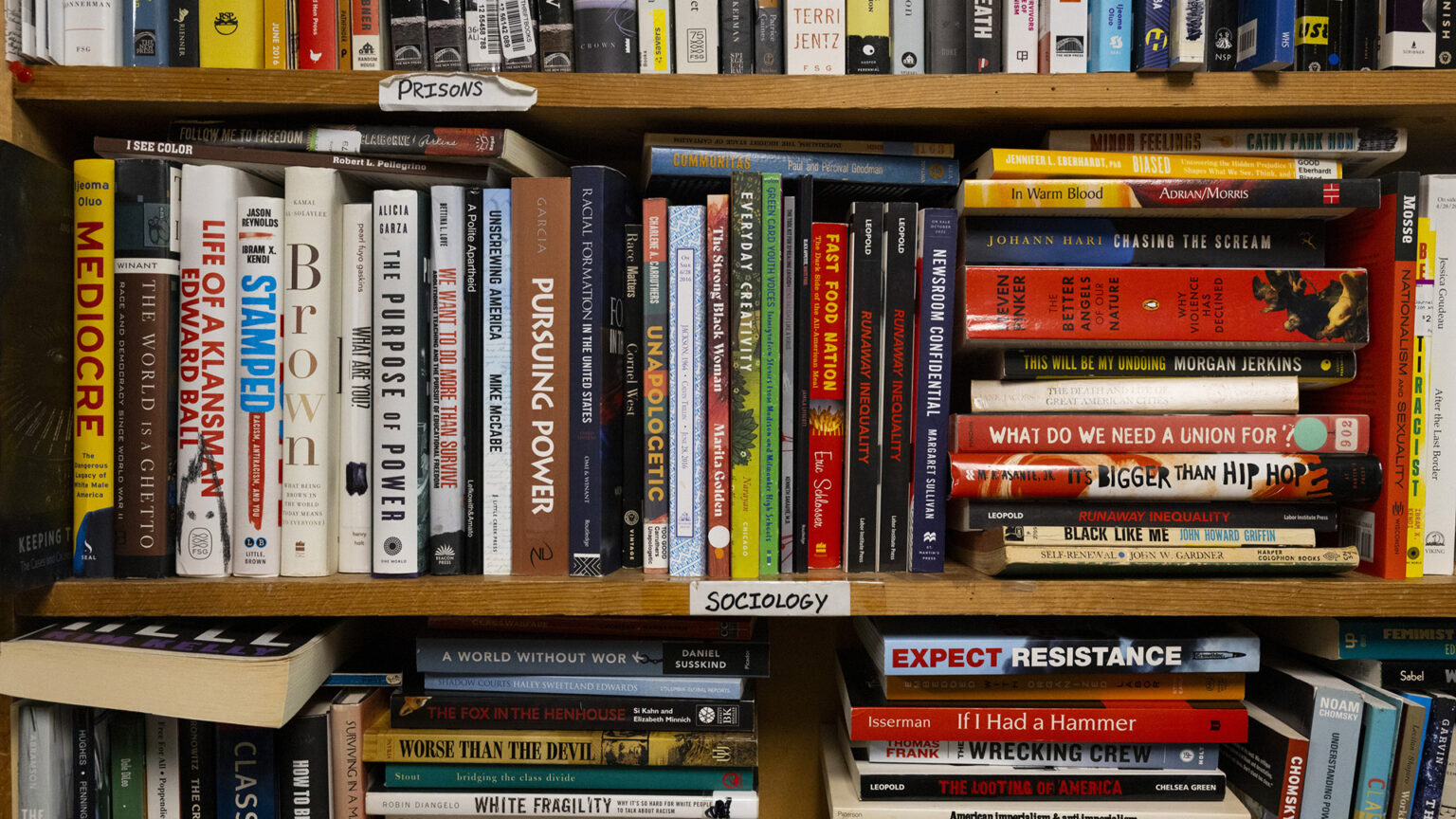 A wood bookcase with paper labels marked Prisons and Sociology taped to the edge of two shelfs is filled with books stacked vertically and horizontally.