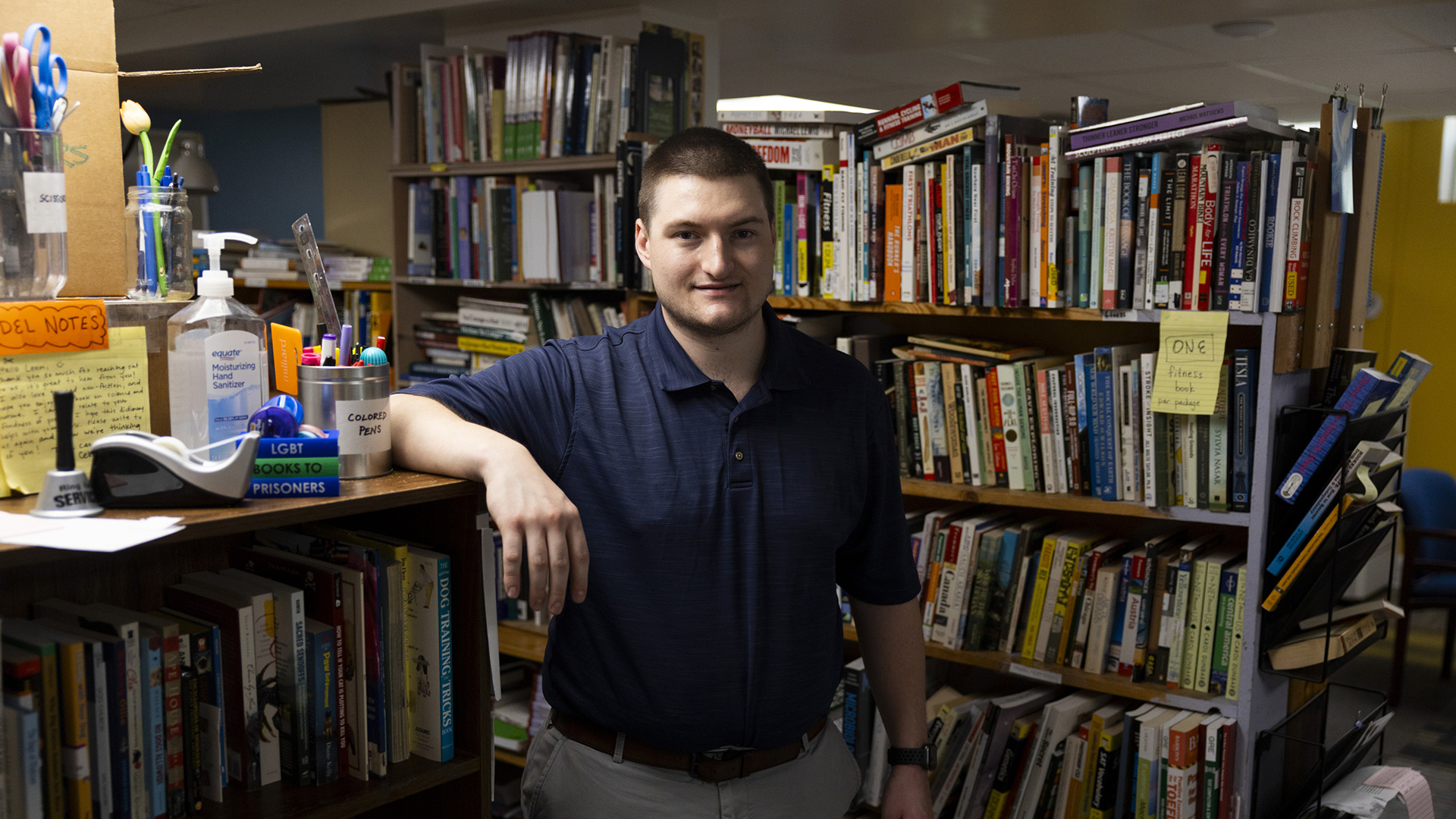 Kyle Wienke poses for a portrait while resting his right forearm on the edge of a bookshelf, with more bookshelves filled with books in the background.