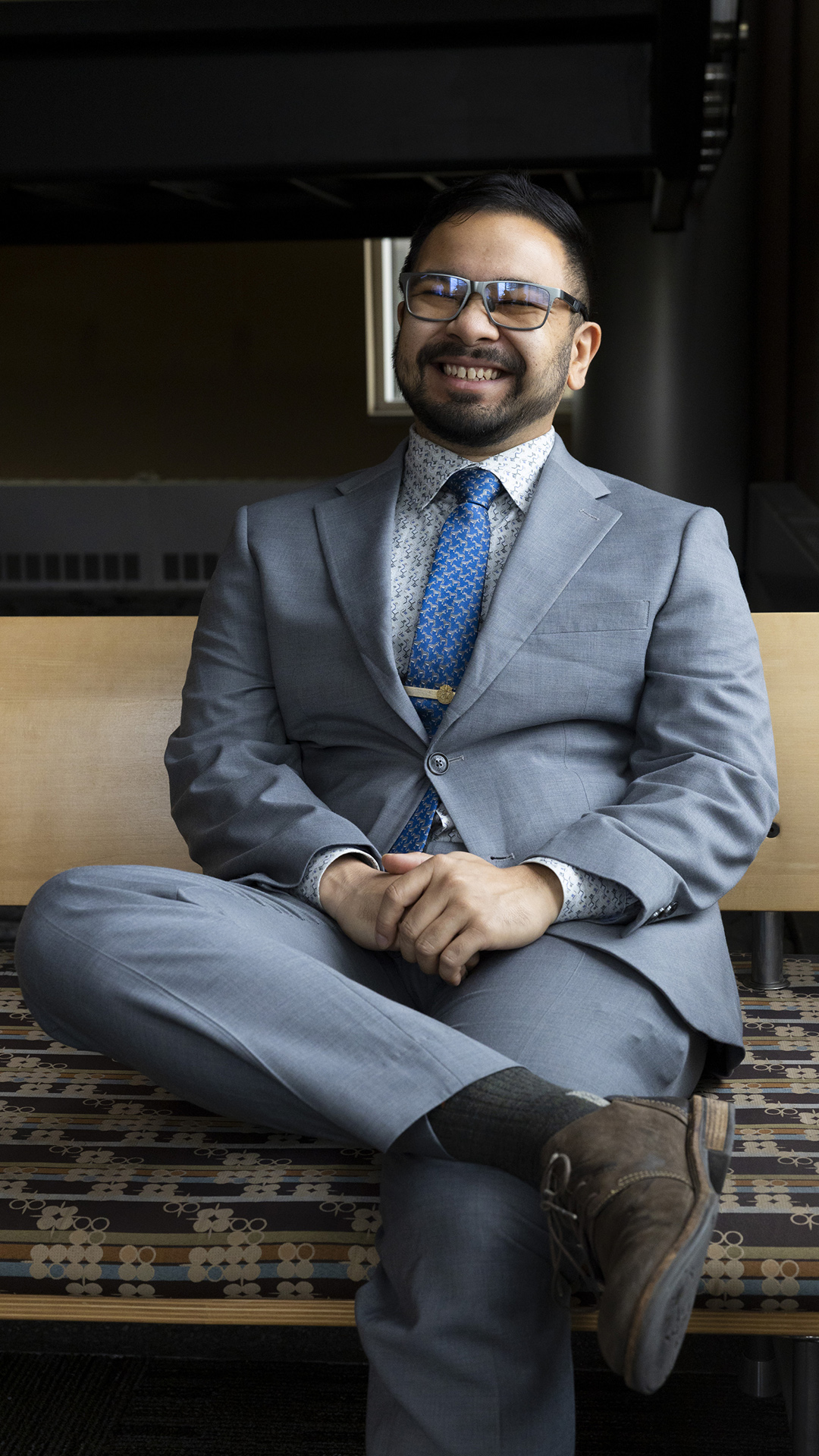 Kai Brito poses for a photo while sitting with legs crossed on a bench with an upholstered seat and wood back.