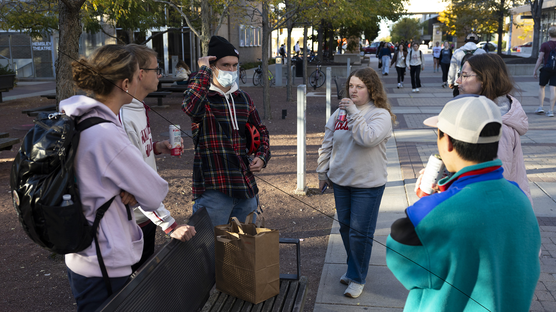 Six people stand in a circle around one end of a metal park bench in a plaza with a paved area and garden area with a gravel surface, with people walking among trees and buildings in the background.