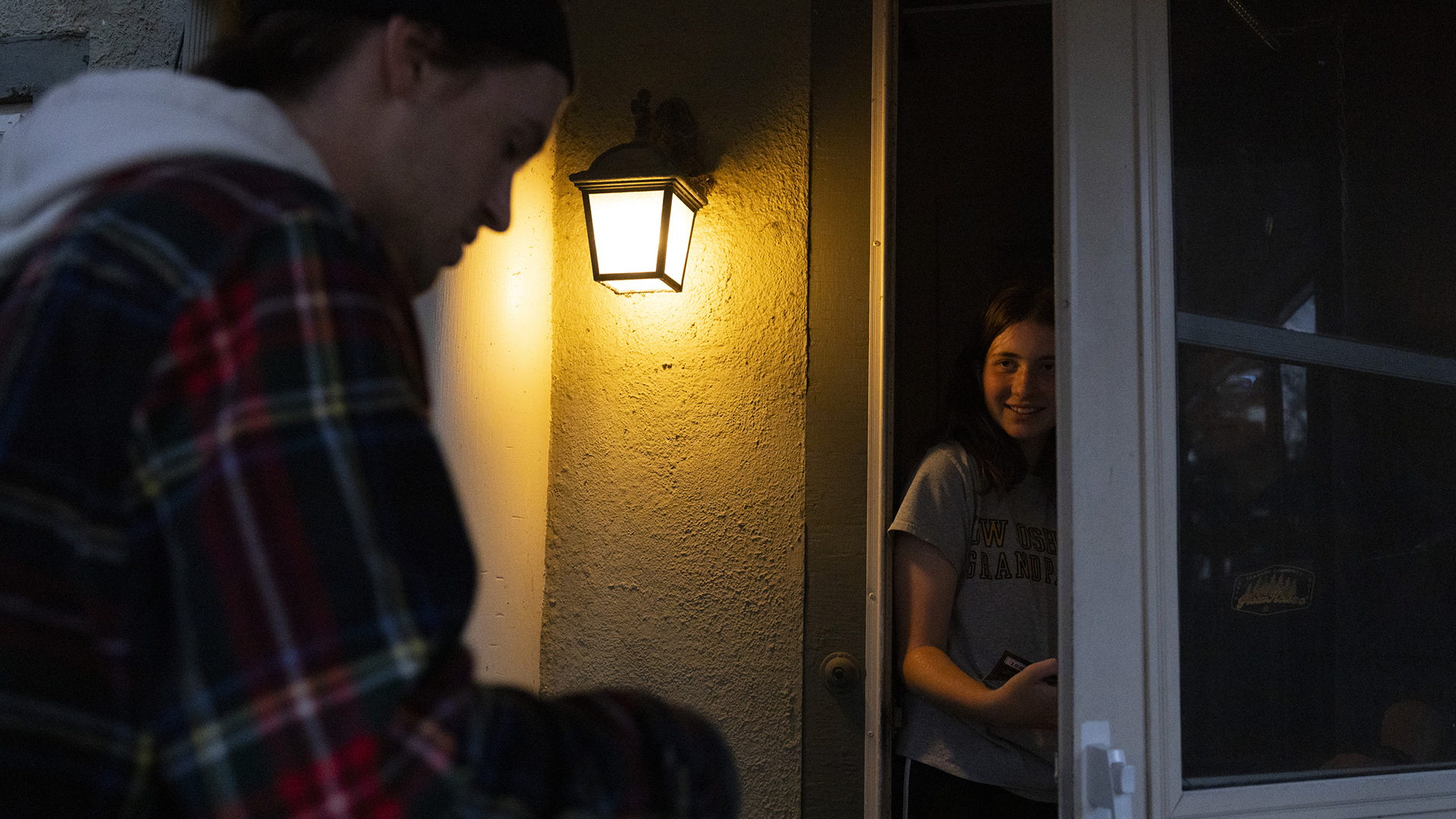 Ty Schanhofer, out-of-focus while standing in the foreground, faces another person standing behind an open screen door to a building with a stucco surface and illuminated outdoor lamp.