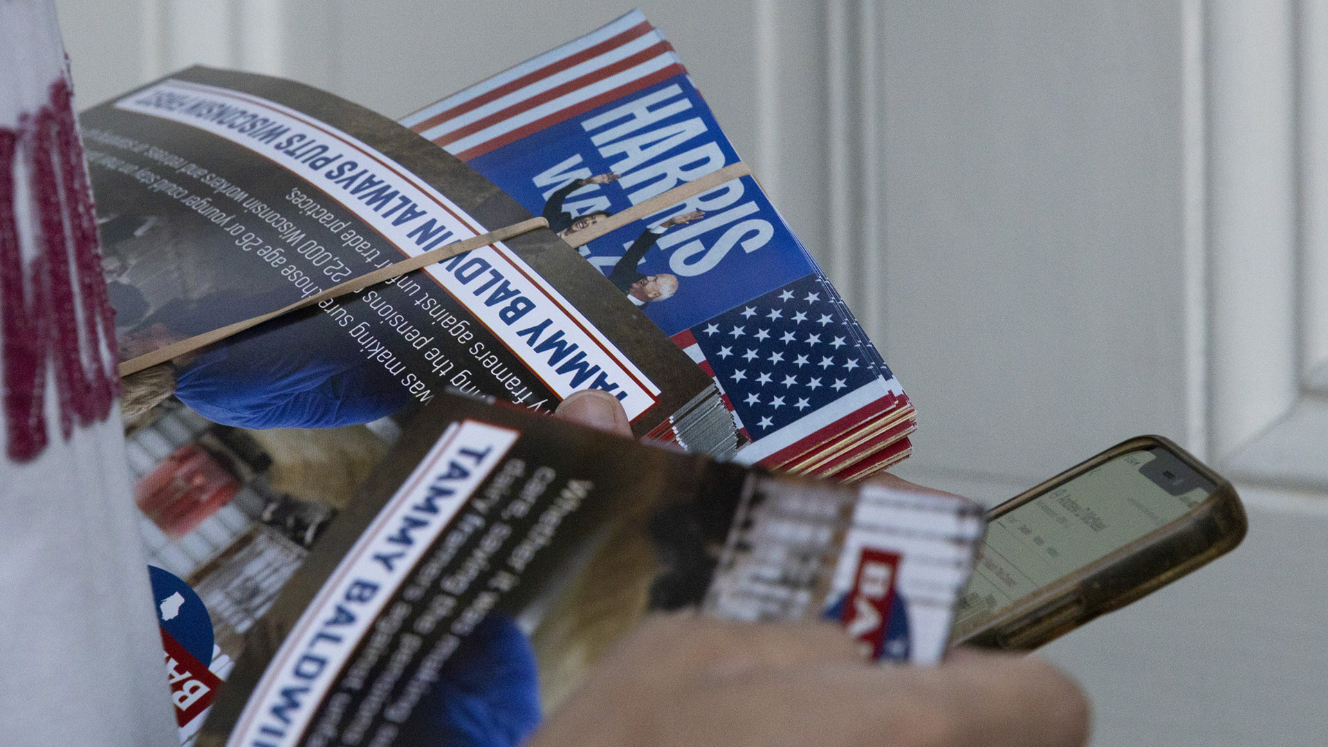 A pair of hands holds a smart phone and multiple stacks of campaign literature held together with rubber bands in front of a closed door.