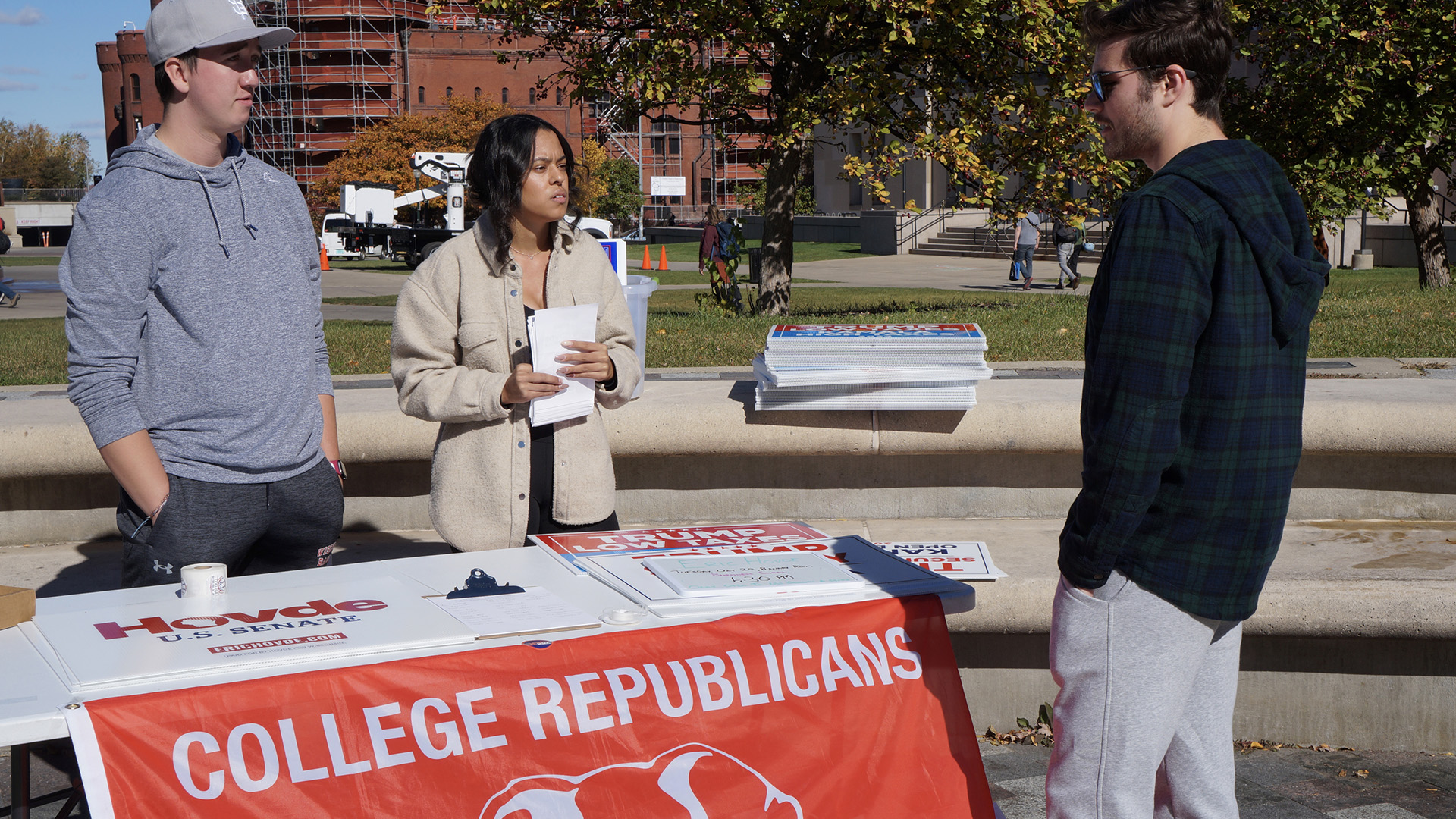 Thomas Pyle and Korina Thoms stand behind a folding table with a flag with the words "College Republicans" draped over its front and with a clipboard and multiple stacks of campaign signs on its surface and talk to another person facing them, with another stack of campaign signs stacked on the upper level of a concrete landscaping bench behind them, and with pedestrians, trees and multiple buildings, one with construction scaffolding around it, in the background.