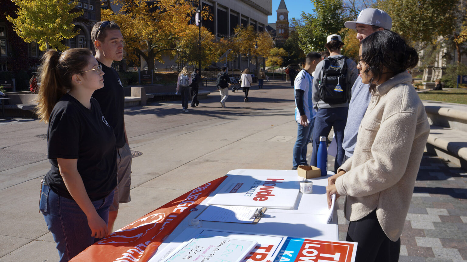 Thomas Pyle and Korina Thoms stand behind a folding table with a flag with the words College Republicans draped over its front and with a clipboard and multiple stacks of campaign signs on its surface and speak with two other people on the other side, with pedestrians, trees and multiple buildings in the background.