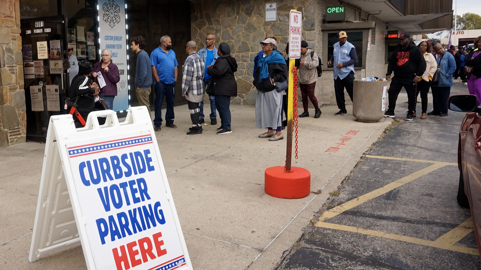 People stand in line outside the entrance to a masonry building, with illuminated neon lights with the words "Open" and "Closed" in place over drive-thru windows, with a sandwich board sign reading "Curbside Voter Parking Here" standing to the side.