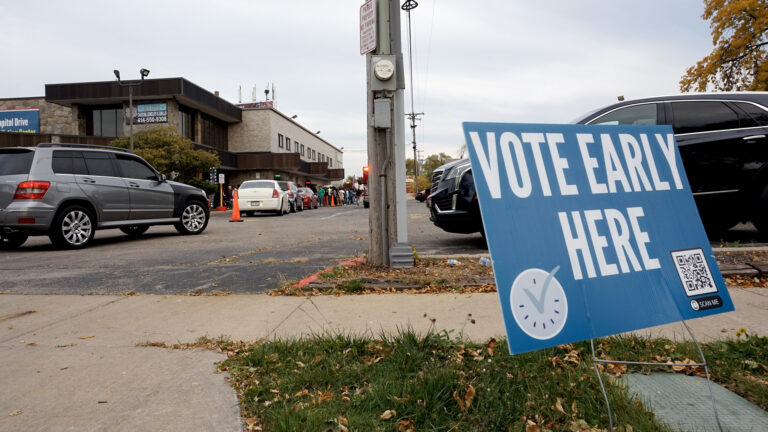 A political yard sign with the words Vote Early Here, a clock graphic with a checkmark and a QR code stands in a grass median between a sidewalk and driveway, with a line of vehicles in a parking lot alongside a multi-story Prairie-style building and a line of people standing outside, with parked vehicles and trees in the background.