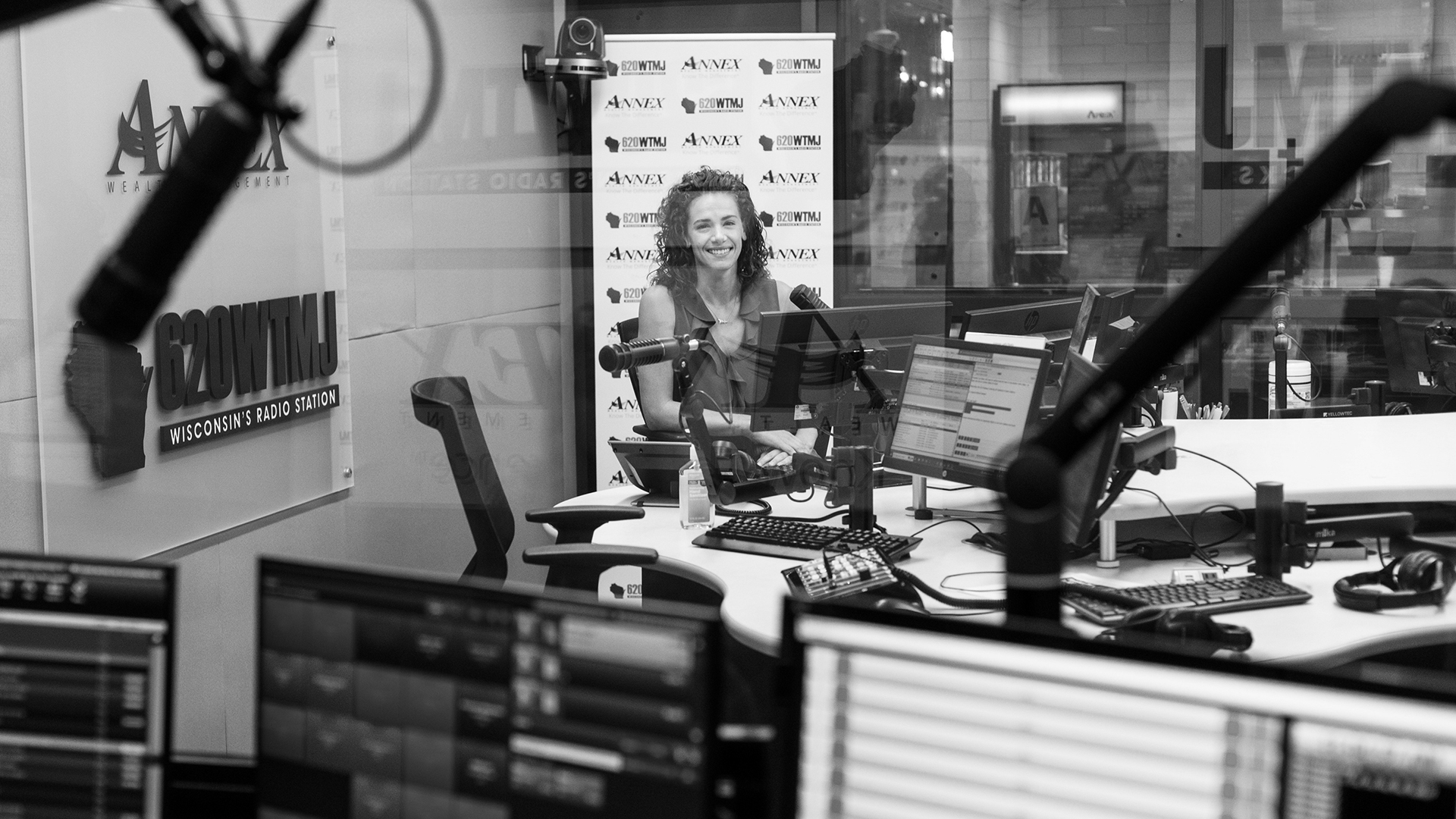 Kristin Brey sits in a radio studio, facing a monitor on one face of a multi-sided table with multiple work stations equipped with monitors, keyboards and mounted microphones, in front of a logo backdrop showing the wordmarks for Annex Wealth Management and 620WTMJ Wisconsin's Radio Station and next to a wall-mounted sign featuring the same two wordmarks, and with out-of-focus monitors in a connected room in the foreground and reflections of the wordmarks visible on the pane of glass separating the two spaces.