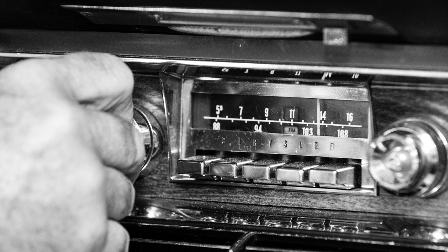 A grayscale photo illustration shows a hand turning the left dial of an AM/FM band car radio with push-button presets and a wood-laminate housing in a Chrysler automobile.