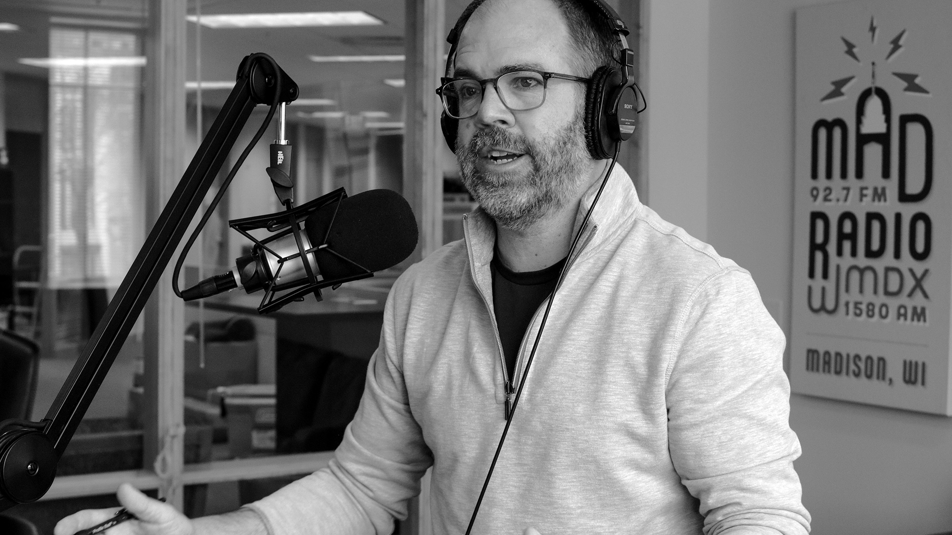 Todd Allbaugh sits and speaks into a microphone mounted to a boom arm in a studio with a glass-paneled wall and a sign showing the Mad Radio word mark with the words "92.7 FM," "WMDX," "1580 AM," and "Madison, WI" in the background.