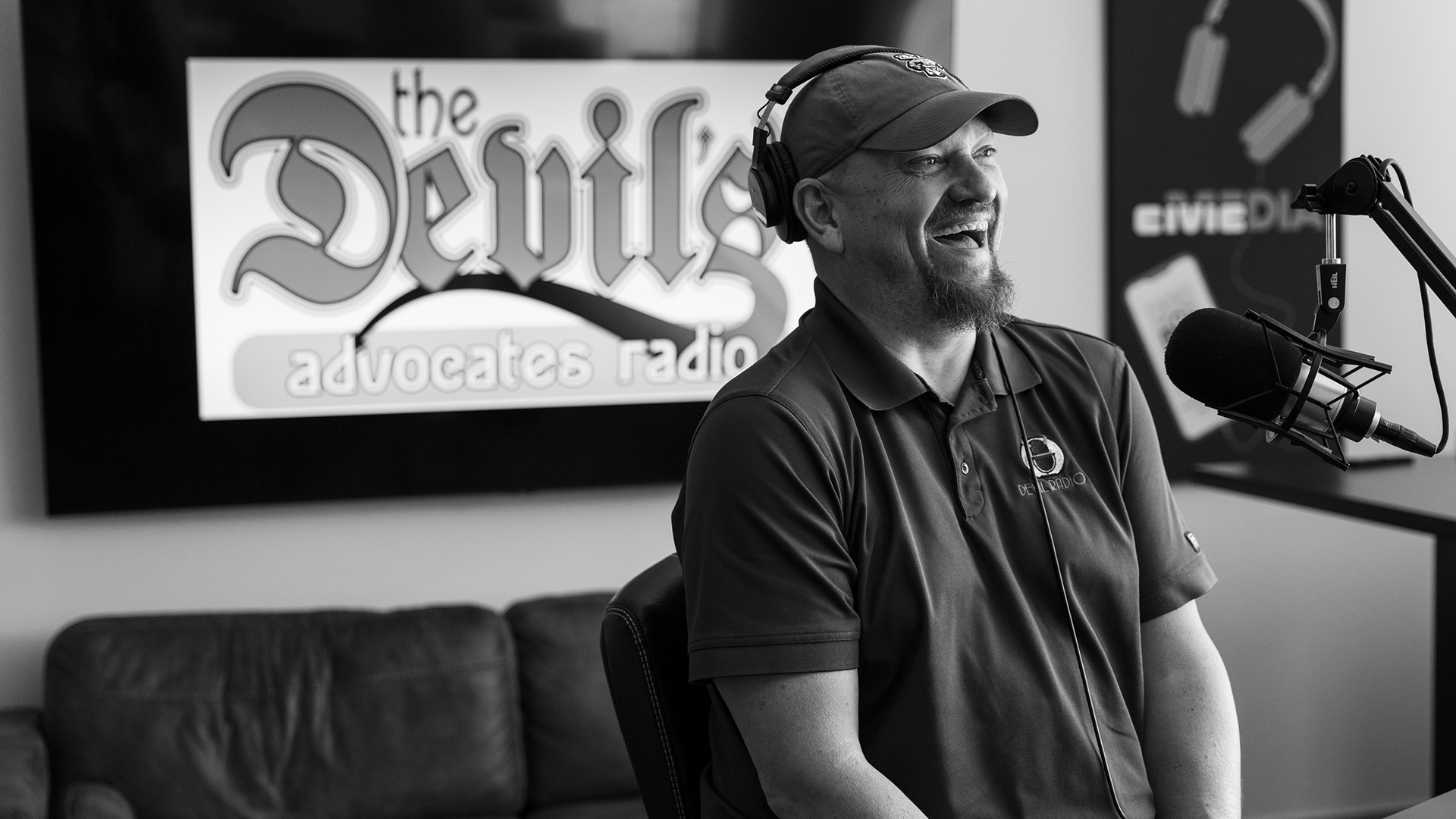 Mike Crute laughs while sitting in front of a microphone mounted to a boom arm, with a leather couch, monitor showing "The Devil's Advocates Radio" wordmark and a sign with the Civic Media wordmark and an illustration of headphones and a smartphone in the background.