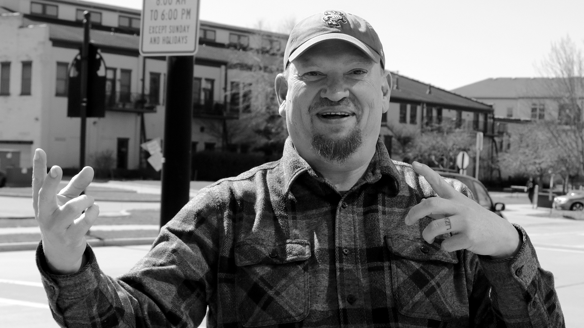 Mike Crute gestures with both hands while standing outside with a road, vehicles, street and traffic light poles, buildings and leafless trees in the background.