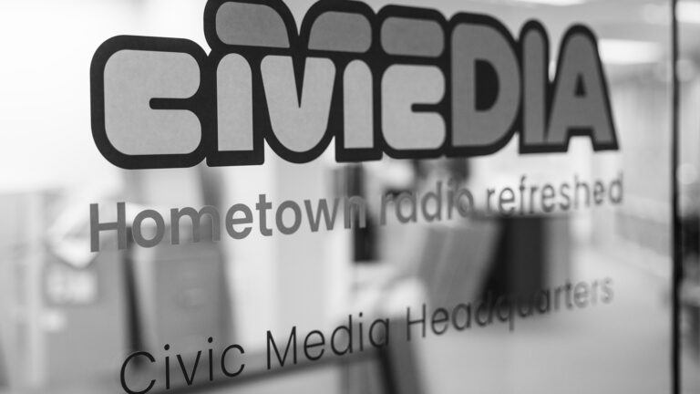 Door decals of Civic Media wordmark and the words Hometown radio refreshed and Civic Media Headquarters are attached to panel of glass, with an out-of-focus office on the other side.