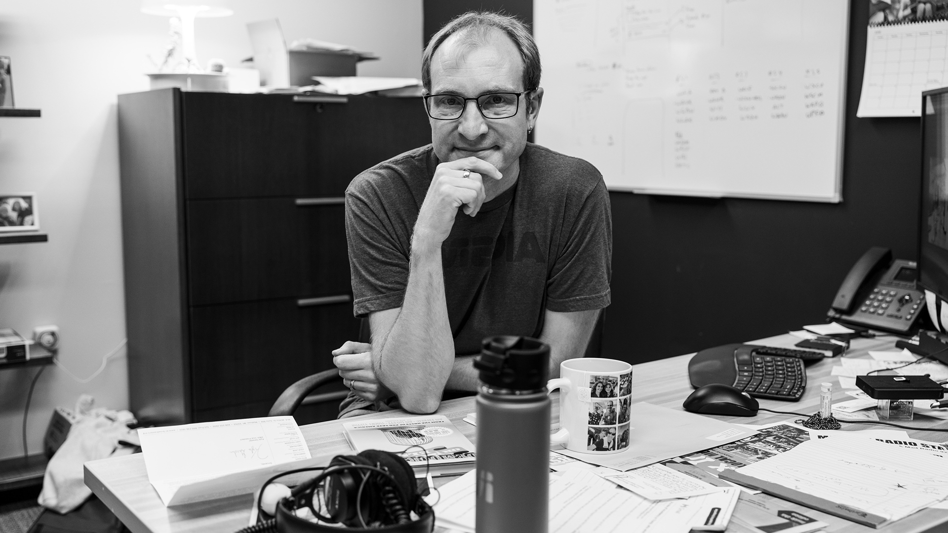 Sage Weil holds his right hand to his chin while sitting jn an office at a desk with papers, a coffee mug and a vacuum-insulated container, keyboard and monitor, phone, headphones, and other items on its surface, with a file cabinet, whiteboard, calendar and other items in the background